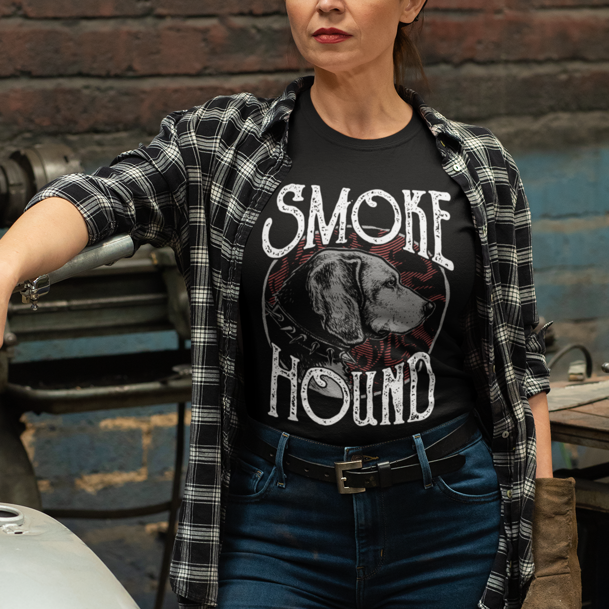 Woman wearing a 'Smoke Hound' t-shirt standing next to a motorcycle in an industrial setting.