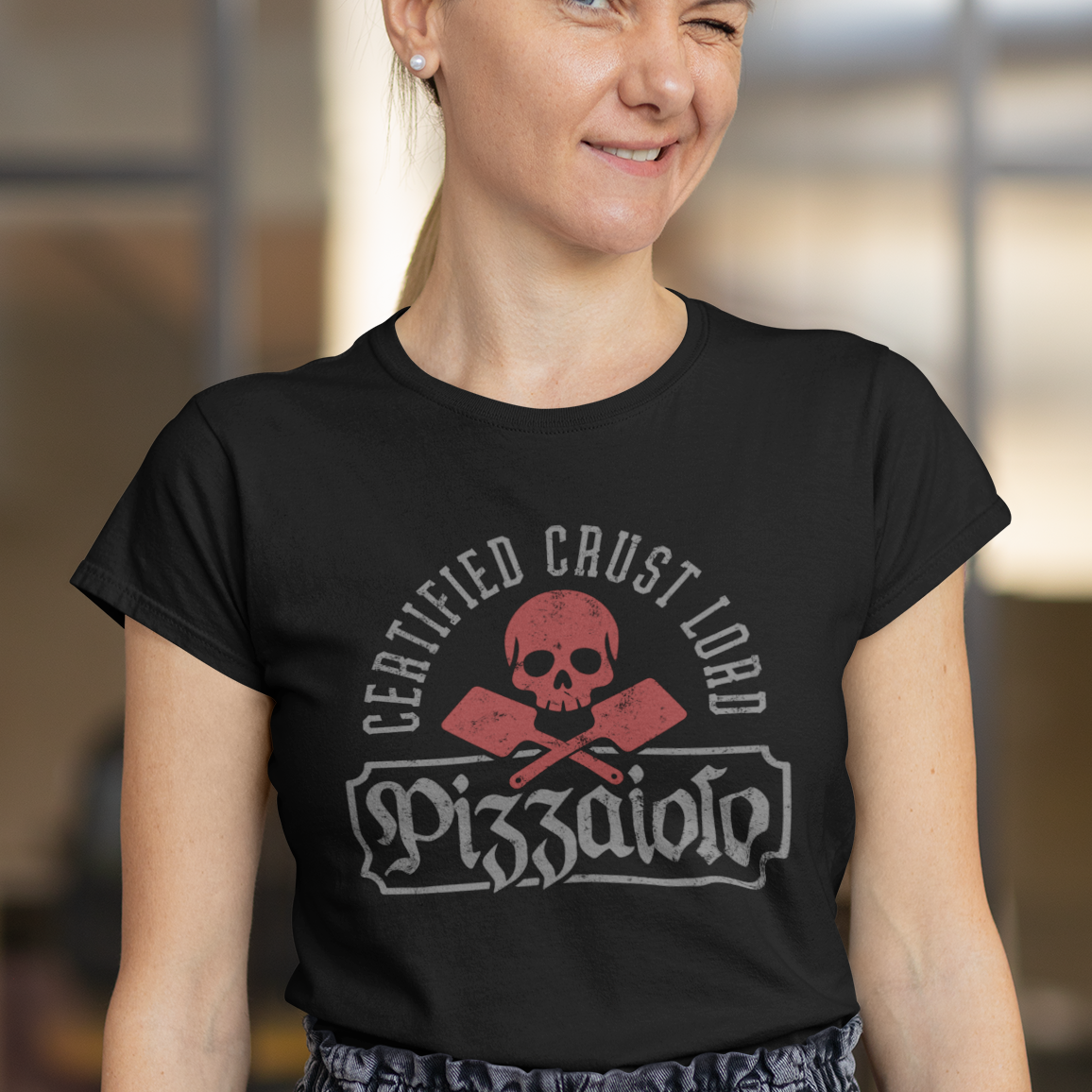 A woman wearing a black short-sleeve t-shirt with a distressed red and white graphic that reads 'CERTIFIED CRUST LORD' and 'Pizzeria 501' around a skull-and-crossbones motif. She is also wearing gray denim jeans, standing in a blurred indoor environment.