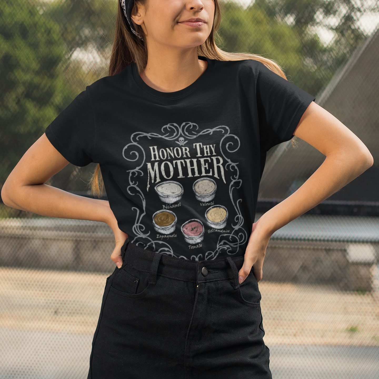 A woman wearing a black graphic t-shirt with the text 'HONOR THY MOTHER' and makeup pans, paired with a black denim mini skirt, posing outdoors.