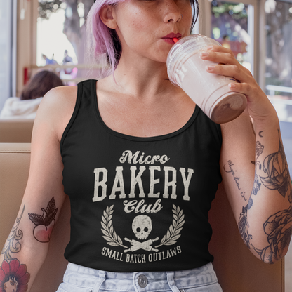 A woman with pink hair sits in a cafe wearing a black tank top with a white graphic that reads 'Micro BAKERY Club' and 'Small Batch Outlaws'. She is also wearing a black baseball cap with a white skull logo and drinking from a clear plastic cup.
