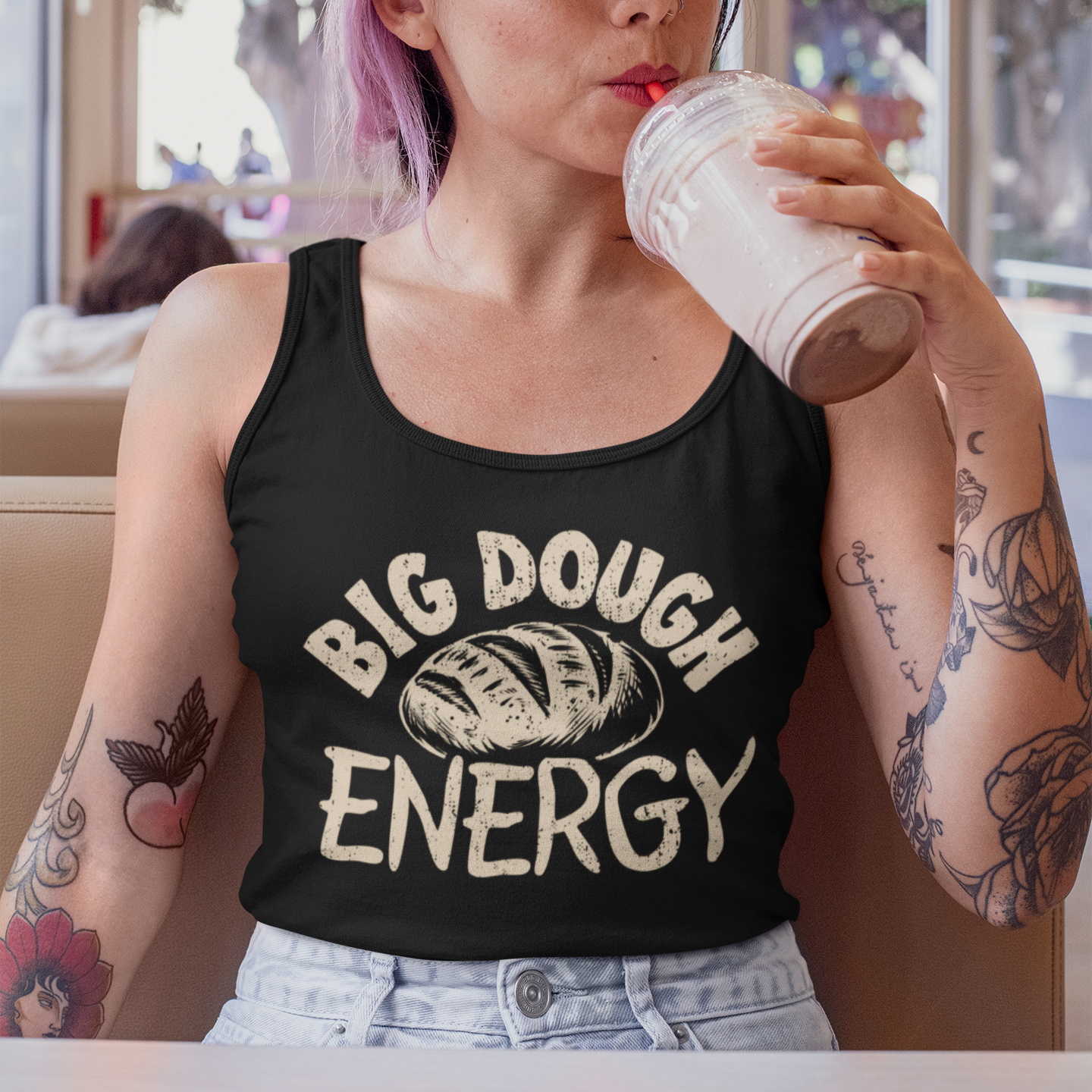 Person wearing a black tank top with 'Big Dough Energy' text, drinking from a paper cup in a casual setting.