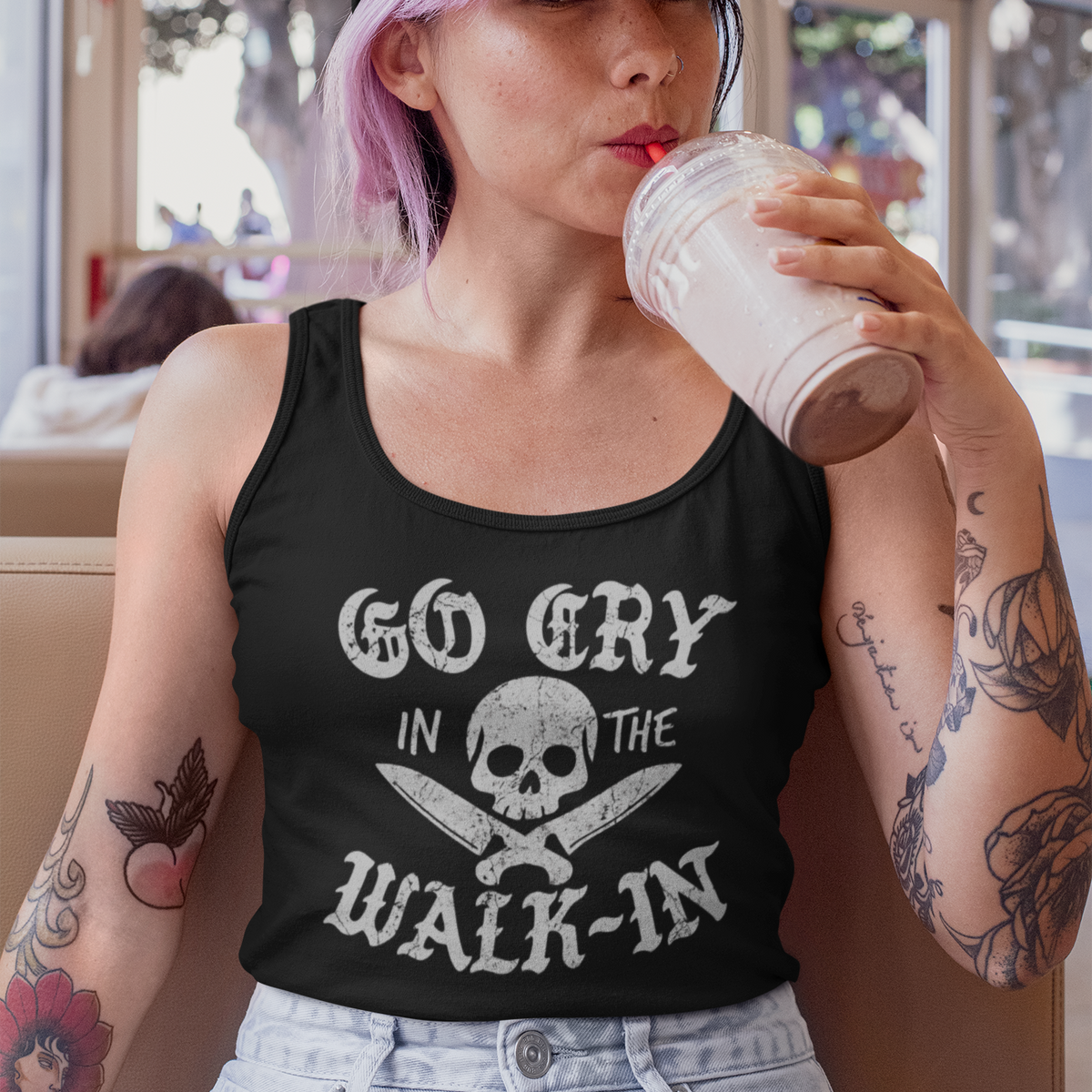 A woman with pink hair wearing a black tank top with a white skull and crossbones graphic that reads 'GO CRY IN THE WALK-IN' and a black baseball cap, sitting in a cafe and drinking from a plastic cup.