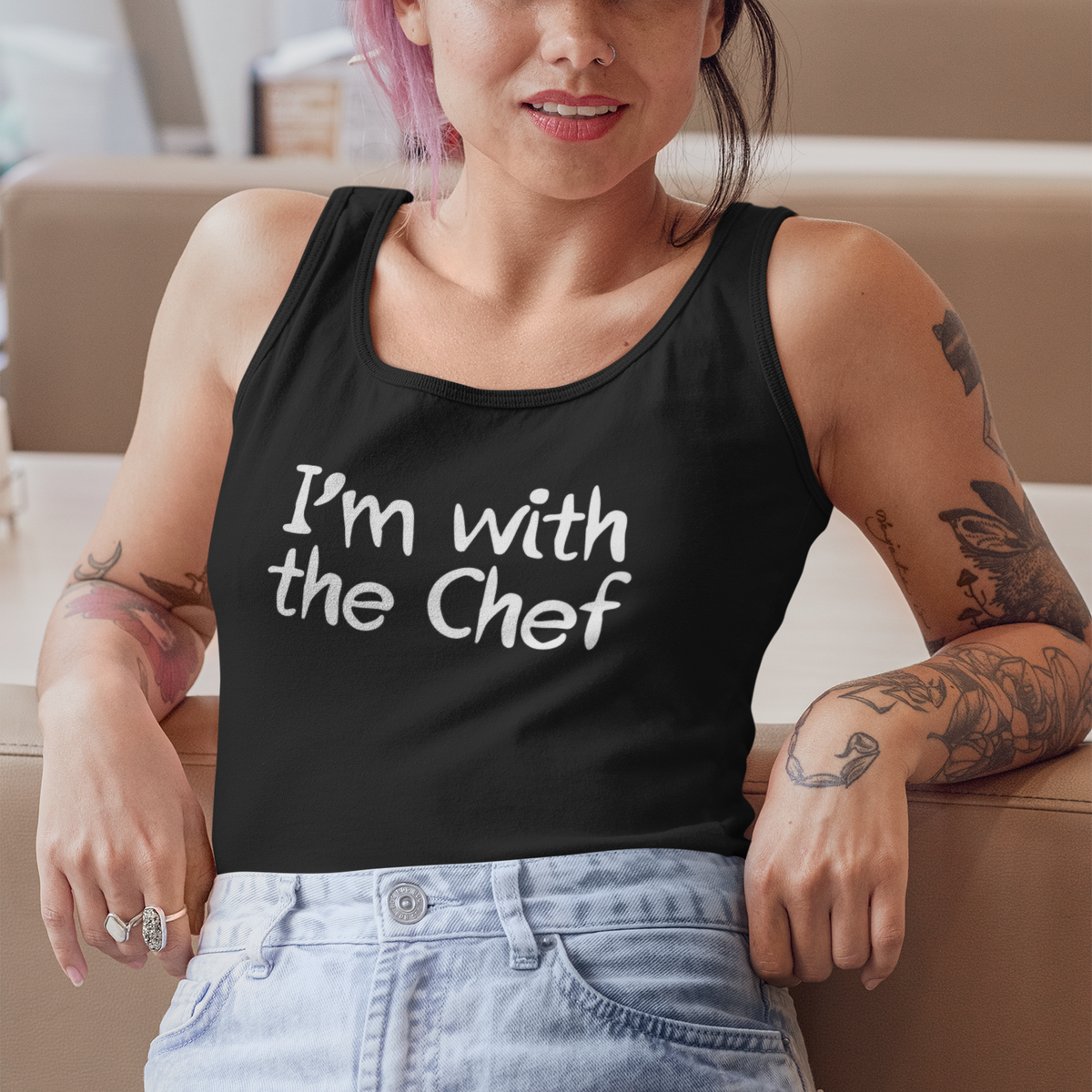 A woman with pink hair, tattoos, and a black cap leans against a beige counter. She is wearing a black tank top with the white text 'I'm with the Chef' and light blue jeans.