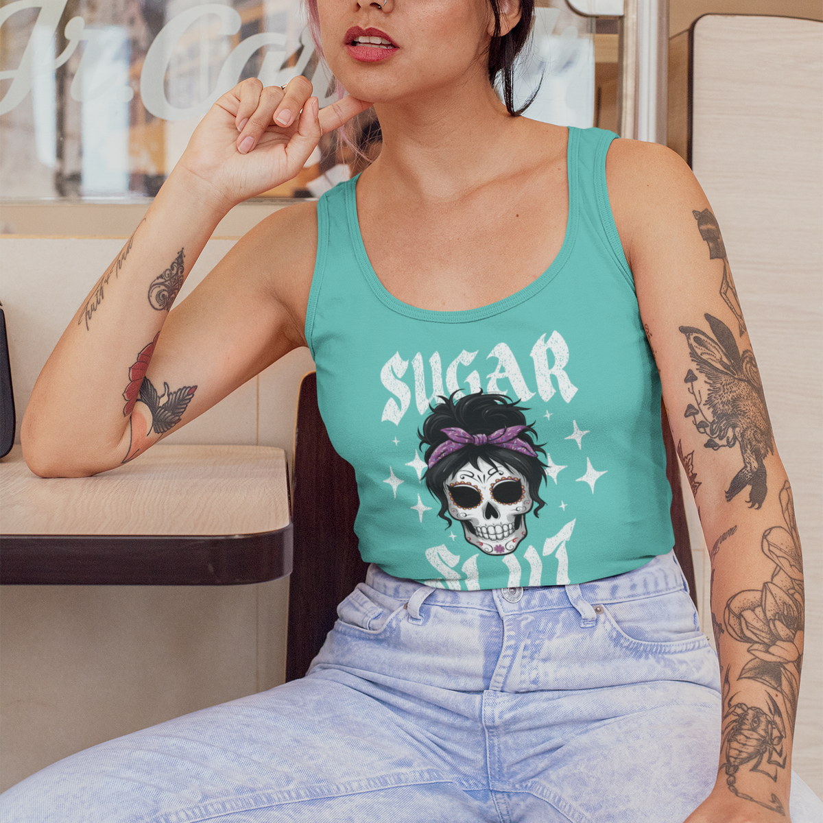 A woman wearing a teal tank top with a sugar skull graphic and the text 'SUGAR SHIT' sits in a diner-style setting. She is also wearing a black baseball cap and light-wash jeans.