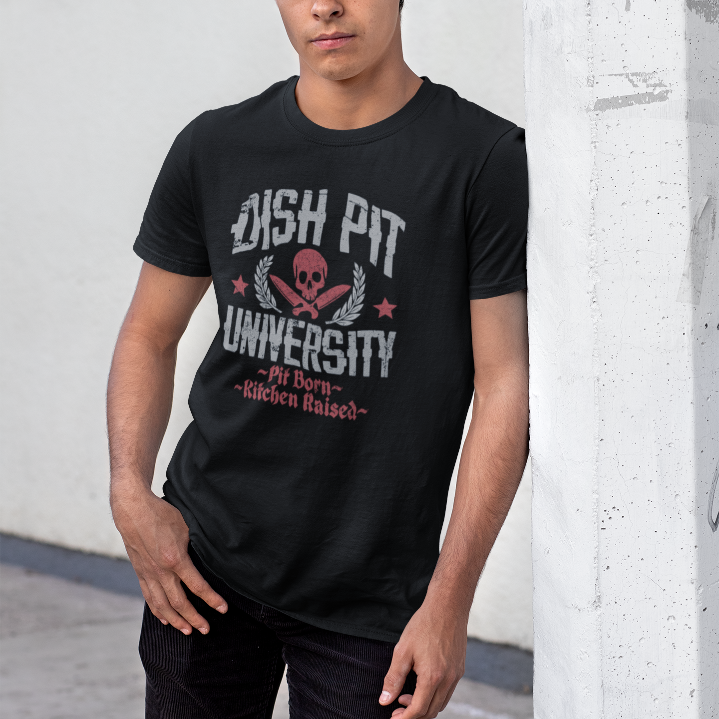 A man wearing a black t-shirt with a 'DISH PIT UNIVERSITY' graphic and a black baseball cap with 'BOTH COM' printed on it, standing against a concrete pillar.