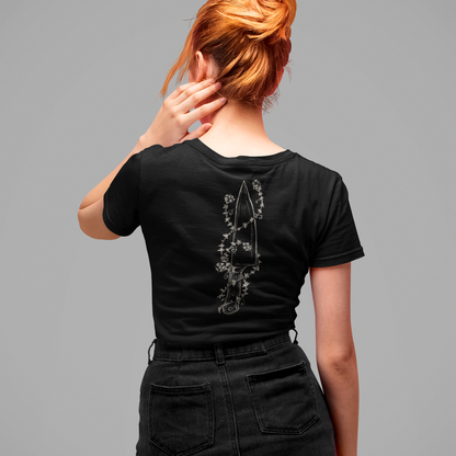 A woman with red hair in a bun is shown from behind wearing a black t-shirt with a silver-toned floral dagger graphic on the back, paired with a black denim skirt, against a solid gray background.
