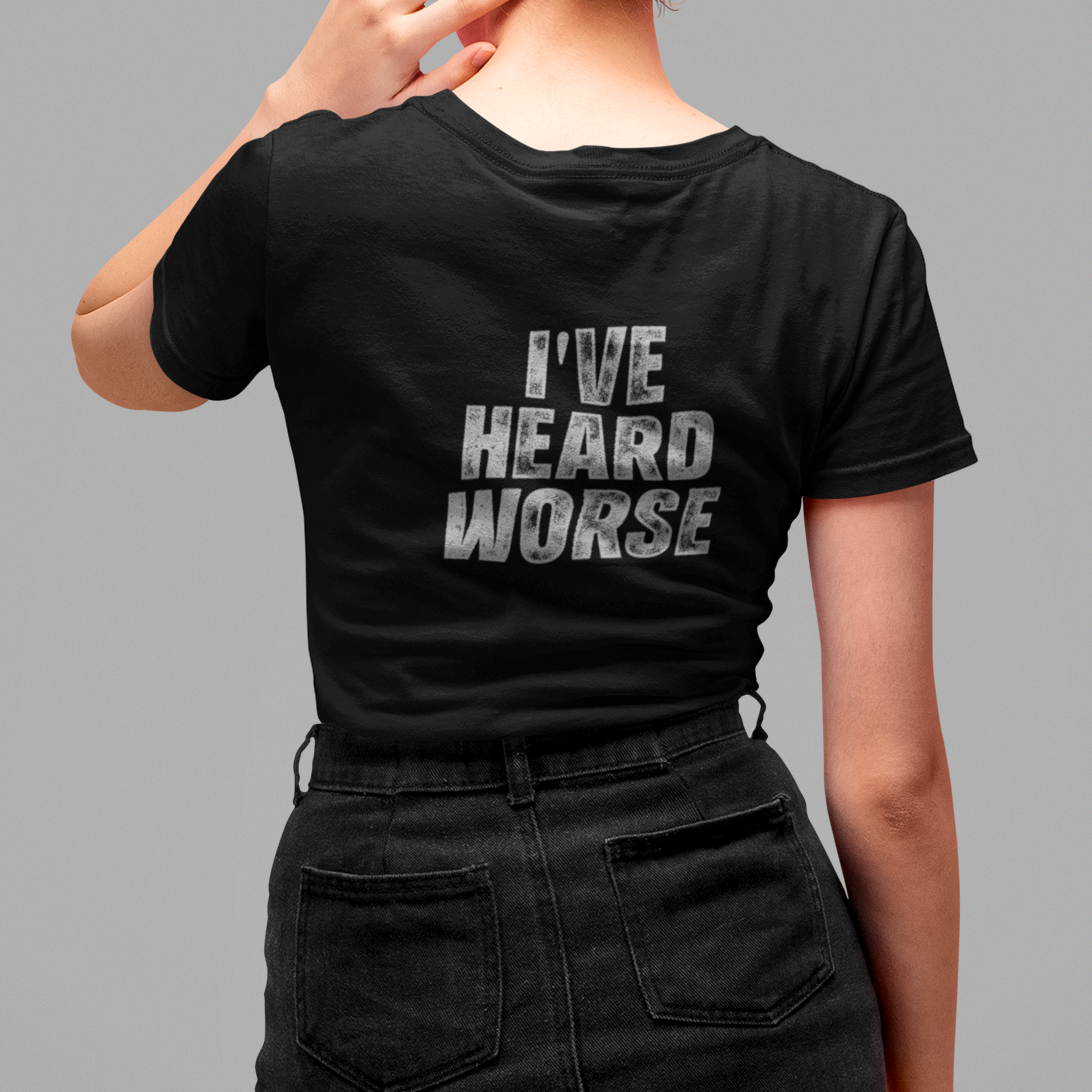 A woman with red hair in a bun is shown from the back wearing a black t-shirt with the text 'I'VE HEARD WORSE' printed in distressed white letters on the back, paired with a black denim skirt, against a solid gray background.