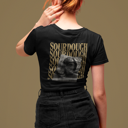 A woman with red hair in a bun is shown from behind wearing a black t-shirt with a large graphic print on the back. The print features the word 'SOURDOUGH' repeated vertically in a gold-toned textured font, overlaid with a grayscale photo of a sourdough bread crust.