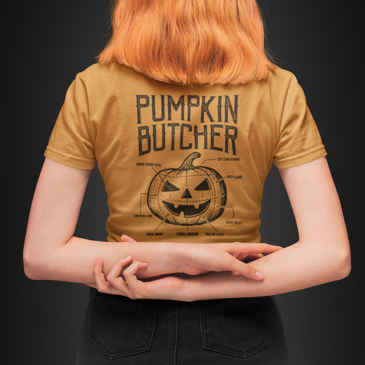 A person with bright orange hair is shown from behind wearing a mustard yellow graphic t-shirt with 'PUMPKIN BUTCHER' and a pumpkin illustration printed on the back, set against a dark studio background.