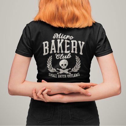 A person viewed from behind wearing a black short-sleeve T-shirt with a large white graphic print reading 'Micro BAKERY Club' and 'Small Batch Outlaws' with a skull and leaf design. The person has bright orange hair and is standing against a plain light gray studio background.