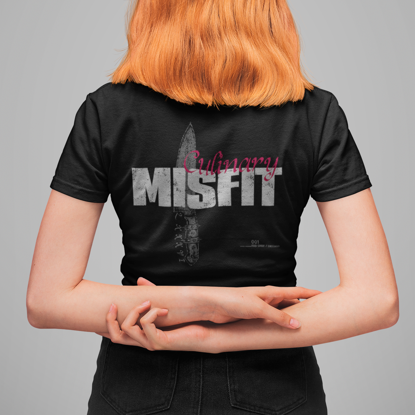 A person shown from behind wearing a black short-sleeve T-shirt with a large distressed white 'MISFIT' graphic and a knife illustration on the back, against a plain gray studio background.