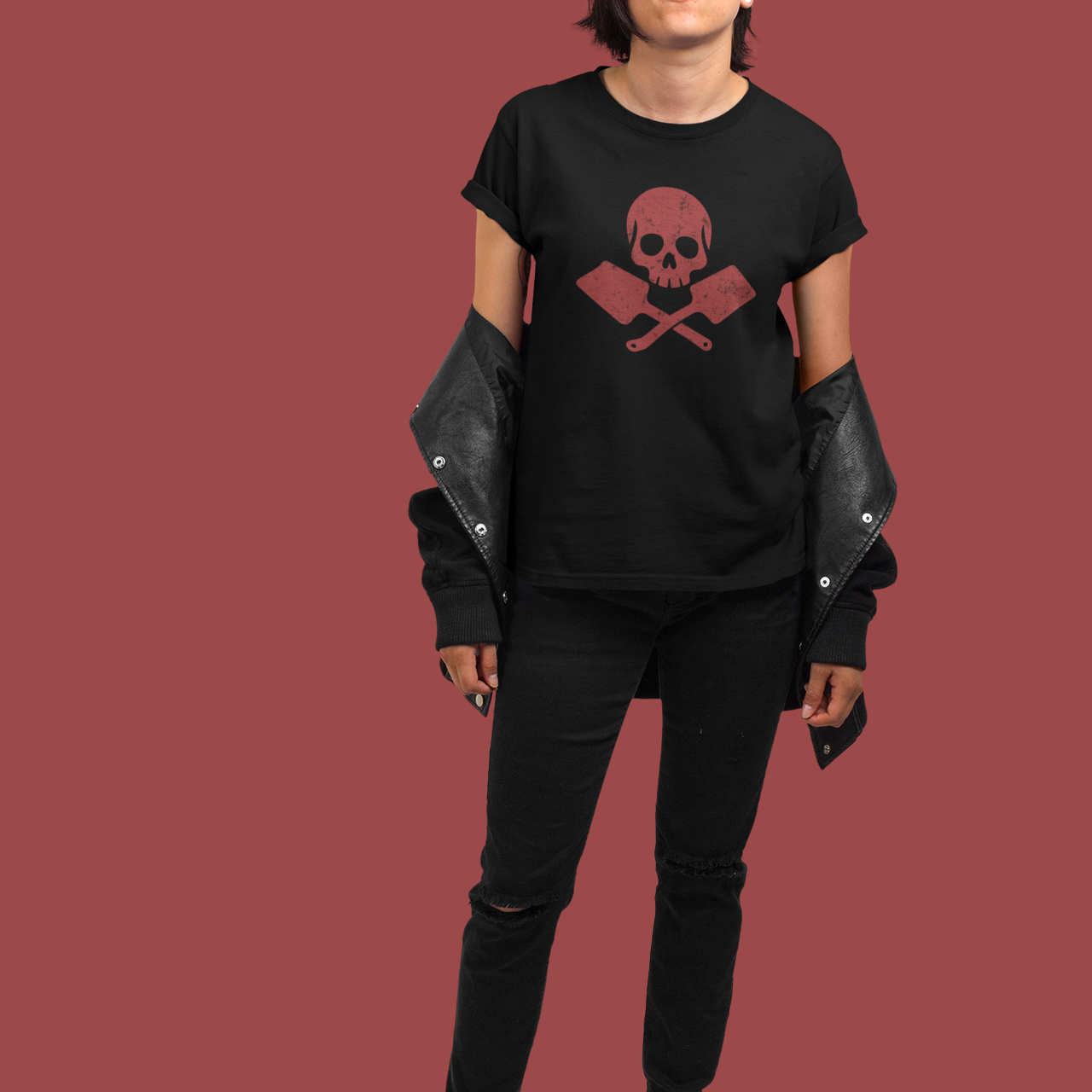 A model stands against a solid dark red background wearing a black t-shirt with a red skull and crossbones graphic, black leather-look jacket sleeves, black jeans, and white sneakers.